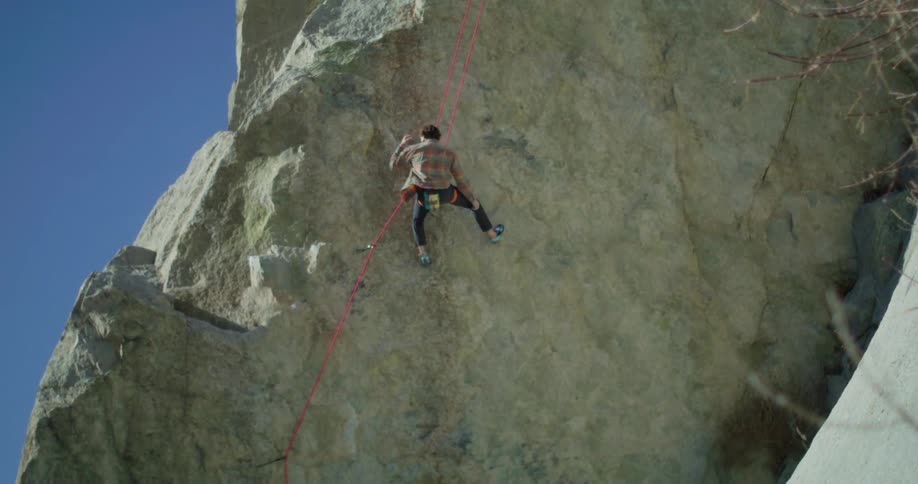 The video shows a wide shot of a person rock climbing on a large palecolored cliff face they are wearing a plaid shirt and dark pants and are attached to a bright red safety rope the sky is a clear deep blue and the watermark filmsupply is centere
