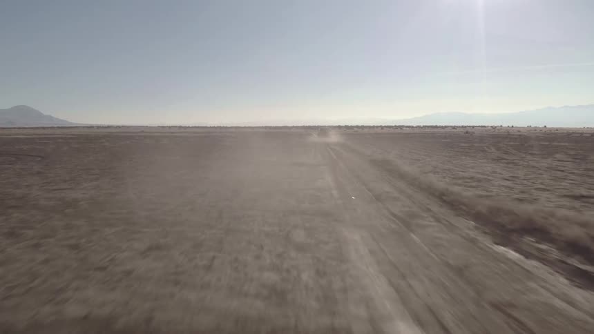 The video is an aerial shot of a vehicle driving along a dusty dirt road in a vast arid landscape the vehicle kicks up a large cloud of dust as it moves away from the camera in the distance a range of mountains is visible against a hazy sky