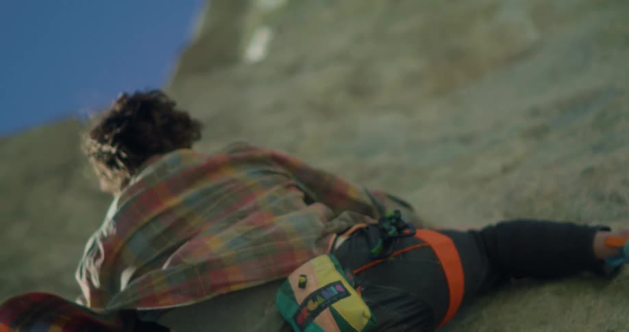 A woman wearing climbing gear scales a steep rock wall under bright daylight