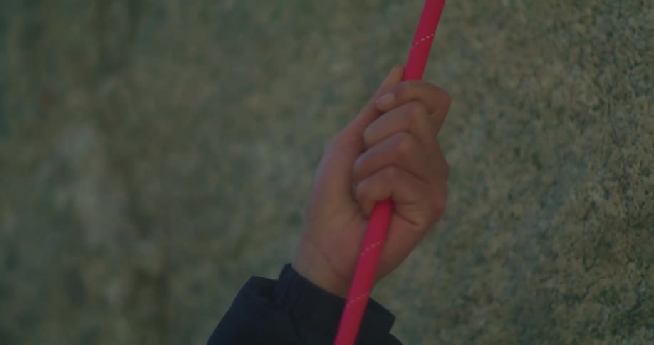 A closeup shot focuses on a persons hand with a dark sleeve gripping a thick red rope the background is a blurry grey and textured surface likely a wall or rock