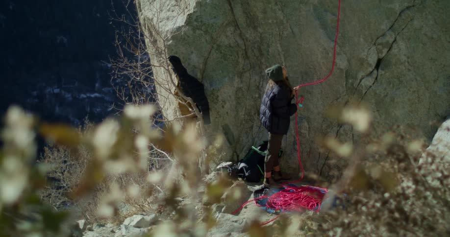 A woman in a black jacket and beanie is standing holding onto a pink rope that is attached to the top of a huge rock