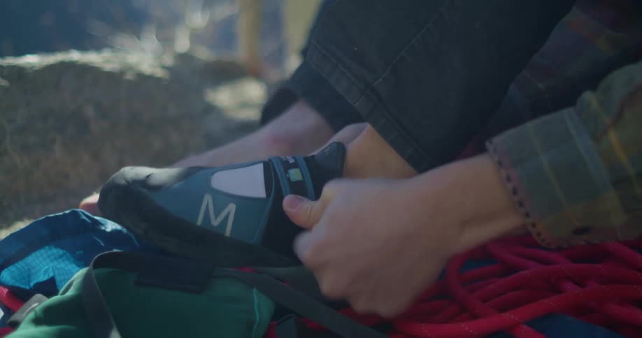 A person is putting on a black and blue climbing shoe with a white m on the side a red climbing rope and other gear are visible on the ground next to them