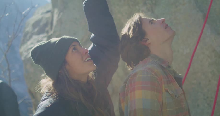A young man and woman are standing outdoors in a mountainous rocky and arid environment the man is wearing a plaid shirt and is looking up while the woman is wearing a dark beanie and is smiling