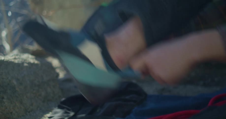 The video is a closeup shot of a person putting on a climbing shoe the person is sitting on a rocky surface and is seen adjusting the strap and heel of the dark blue and black shoe in the blurry background another person and some gear can be seen