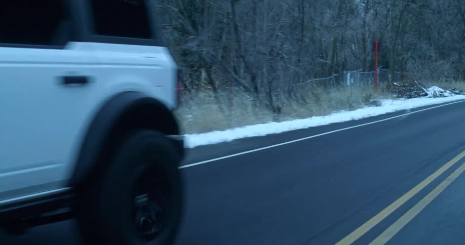 An suv drives on a freeway during winter at midday with snow and leafless trees lining the sides of the road