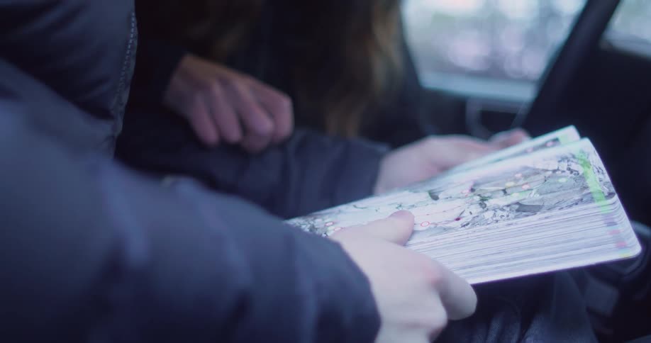 A person in a dark jacket holds an open map inside a vehicle while another sits beside them pointing on the map