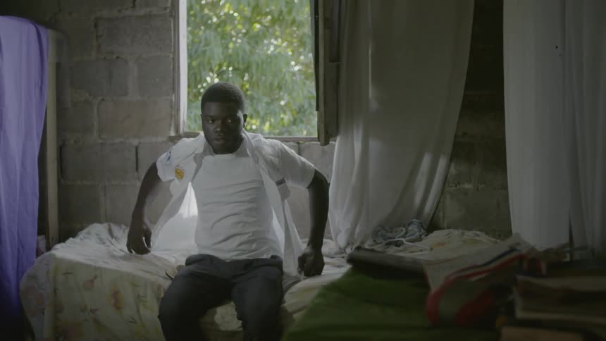 A man sits on a bed and puts on a shirt