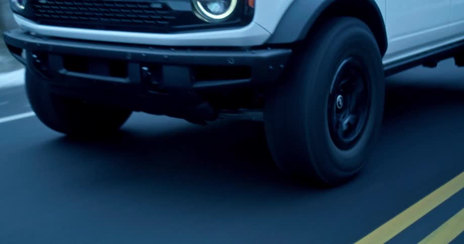 White bronco with headlights on driving down a road the side of the road is covered in snow and the vehicle is moving fast down the paved road