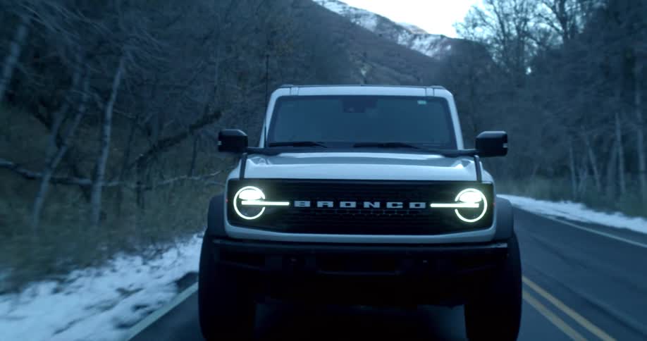 A bronco suv drives on a freeway lined with leafless trees and snow on a winter day