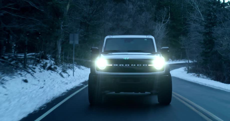 A bronco suv drives on a freeway during winter with the road lined by trees and snow at midday