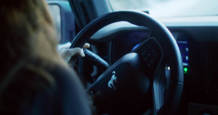 A driver is shown from the shoulder up with their right hand on the steering wheel of a car the logo of a galloping horse is visible on the center of the steering wheel