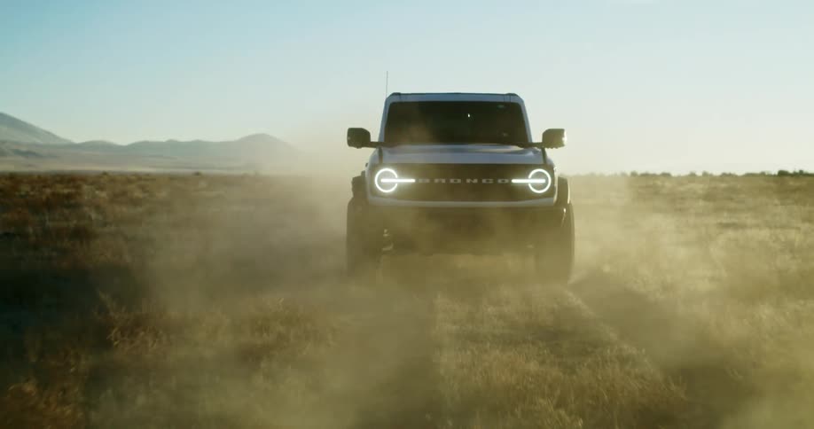 A black ford bronco drives across a dusty open field with mountains in the background at sunset