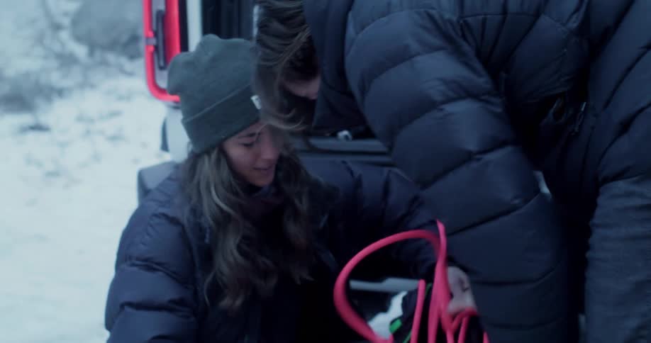 Two women stuff ropes into a bag behind a car both wearing winter jackets at midday
