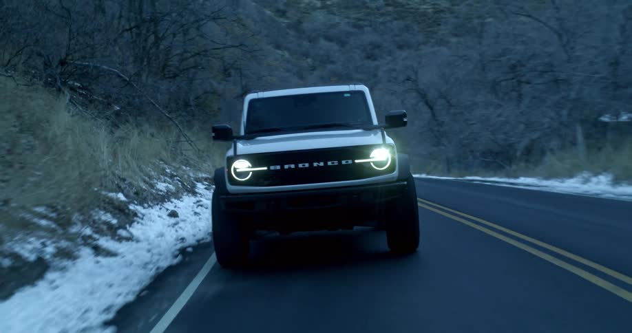 White ford bronco drives along a winding road flanked by snow and bare trees