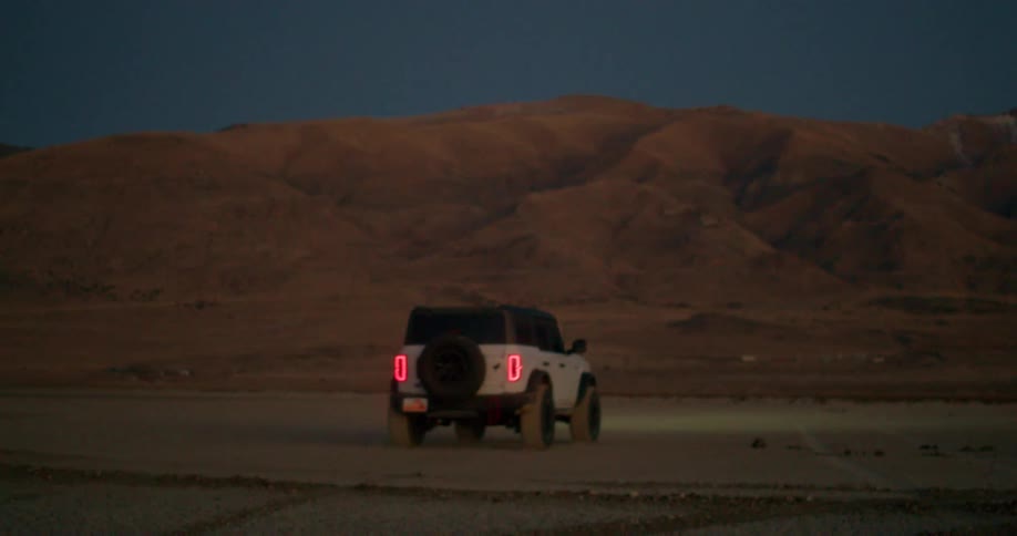 A white ford bronco is driving on a dirt road at dusk the vehicle is kicking up dust and driving toward a large mountain range in the distance