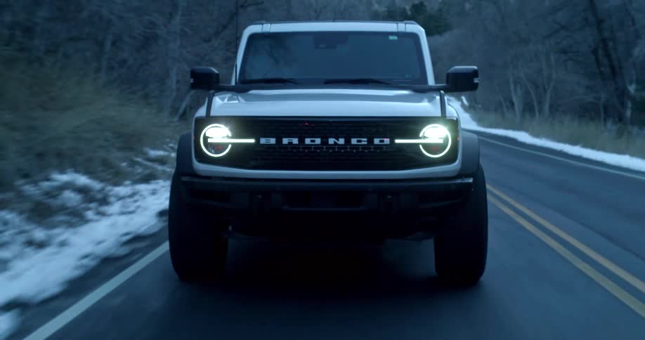 A white ford bronco with headlights on drives down a twolane road bordered by snow and bare trees