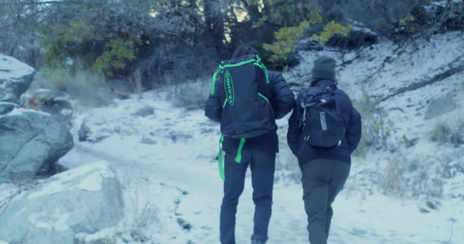 Two hikers wearing winter clothes and backpacks are walking through a snowy rocky landscape one of the backpacks has a neon green trango logo on it