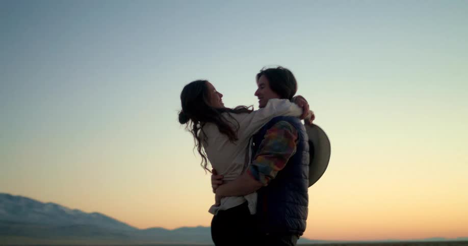 A cowboyhatwearing couple passionately embracing and dancing in an open field against a backdrop of a colorful sunset