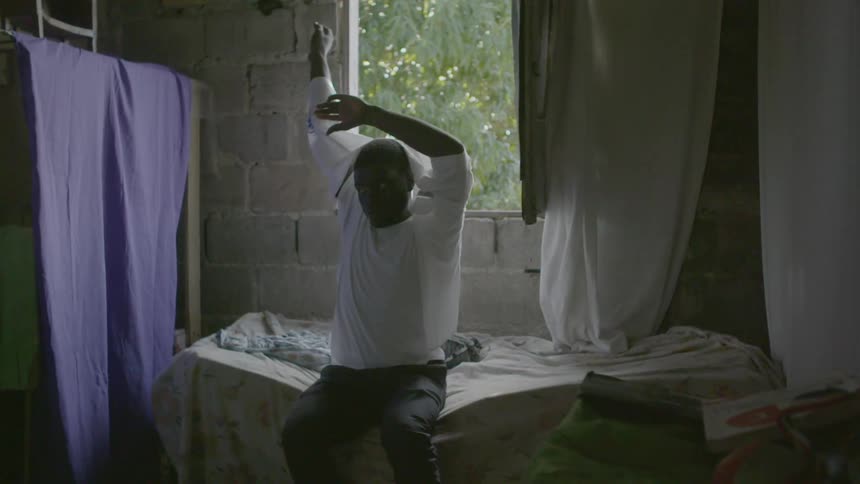 A man sits on a bed and puts on a white shirt