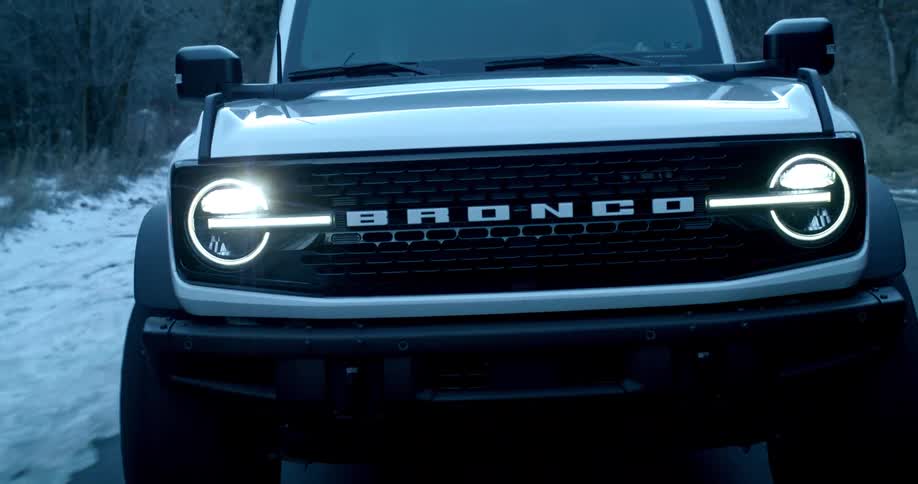 A white ford bronco drives down a snowy forest road with headlights glowing in the cold light
