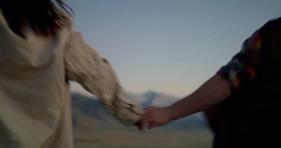 A couple holds hands and walks away from the camera in a wideopen landscape as the sun sets the man is wearing a dark blue vest over a flannel shirt and the woman is wearing a lightcolored jacket