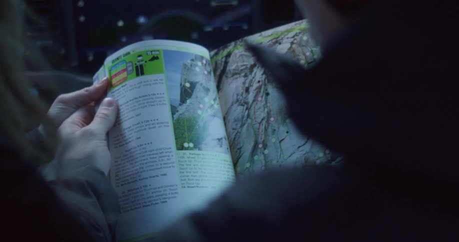 Two people in winter coats inside a car with a touchscreen dashboard looking at a magazine page with a guide to a large mountain
