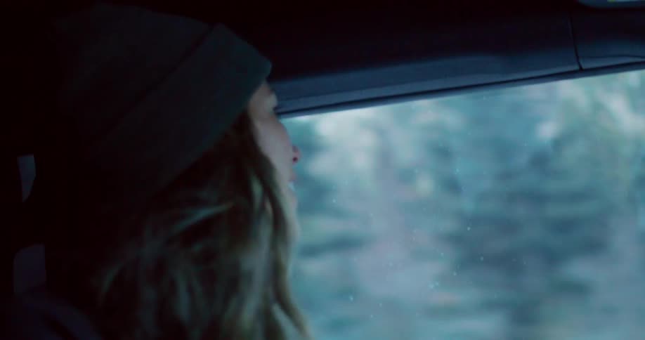 The video shows a closeup of a person with long wavy brown hair and a dark beanie sitting in a vehicle they are looking out the window with a smile and their face is illuminated by light coming from outside the background outside the window is blurr