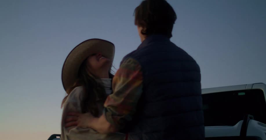 A young couple stands close together at sunset in a wide open landscape the woman wearing a cowboy hat smiles at the man