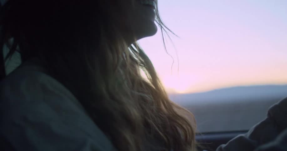 A woman with long hair smiles inside a vehicle as the soft light of sunset casts a warm glow through the window