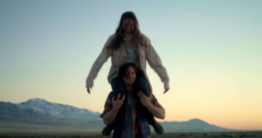 A woman is sitting on a mans shoulders getting a piggyback ride they are in a barren outdoor setting with a mountain range in the distance at sunset