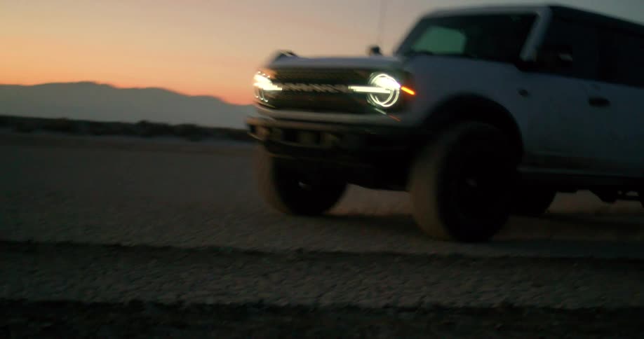 A white ford bronco drives down a dirt road in a desert landscape at sunset the headlights of the truck are on illuminating the road ahead
