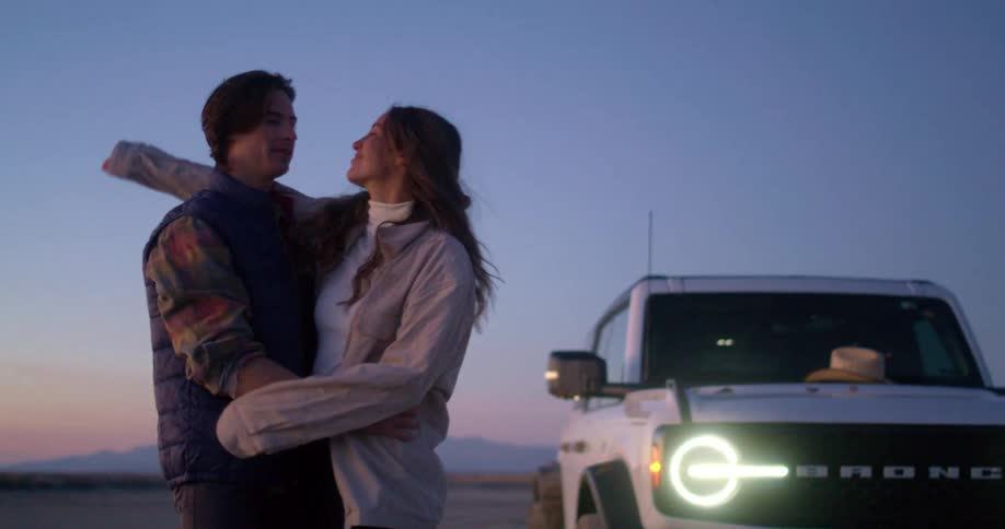 Couple embraces next to a parked ford bronco at sunset