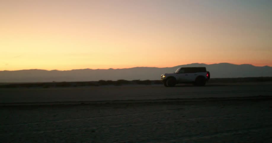 A white suv speeds across a barren desert flat at sunset trailing dust behind it