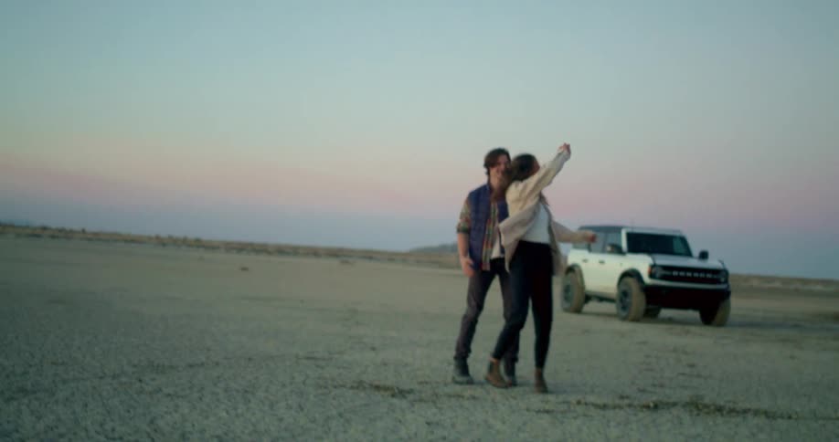 A couple stands in an open area showing affection with a car behind them at midday