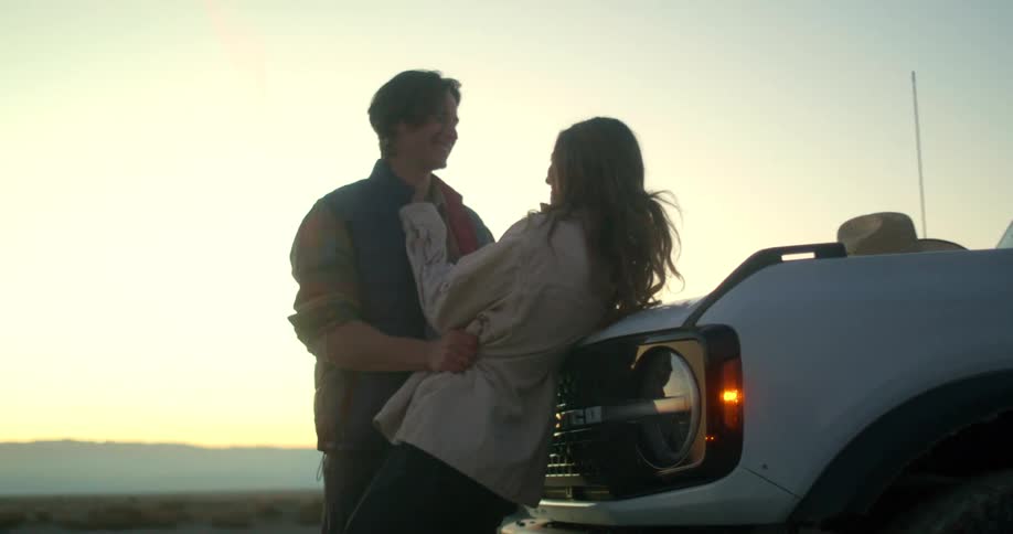 A man kissing a woman who is leaning against the front of a jeep and then she jumps with happiness