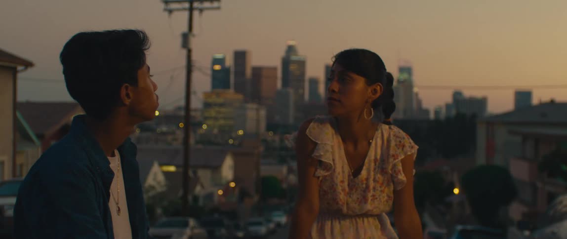 Two young people talk on a sloping street with the city skyline in the background at sunset