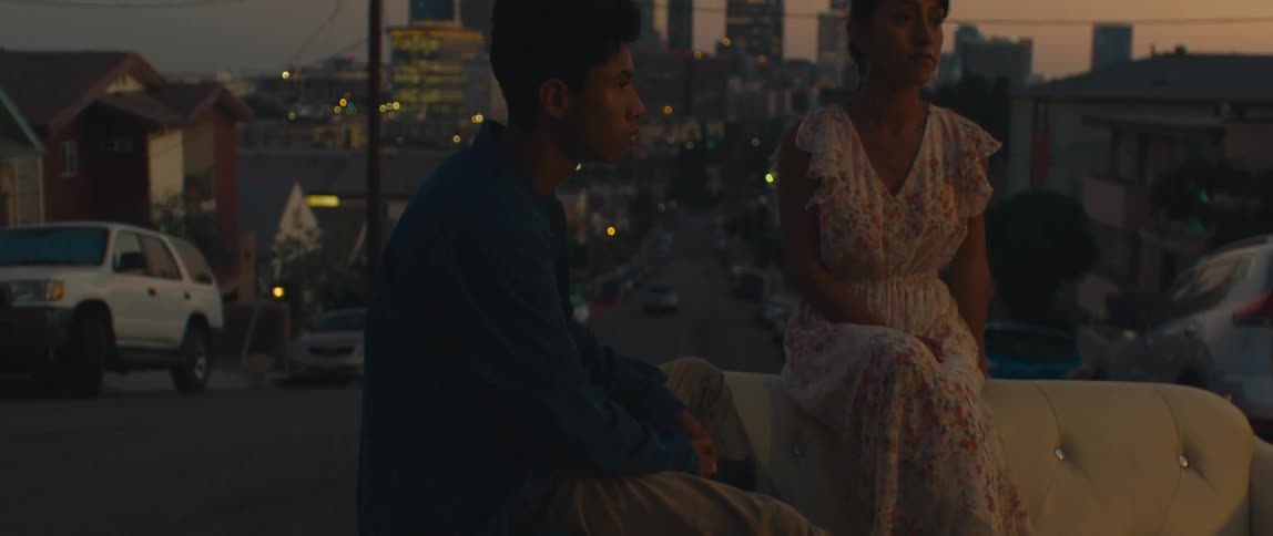 Two young people sit on an outdoor couch with the city lights glowing in the background at dusk