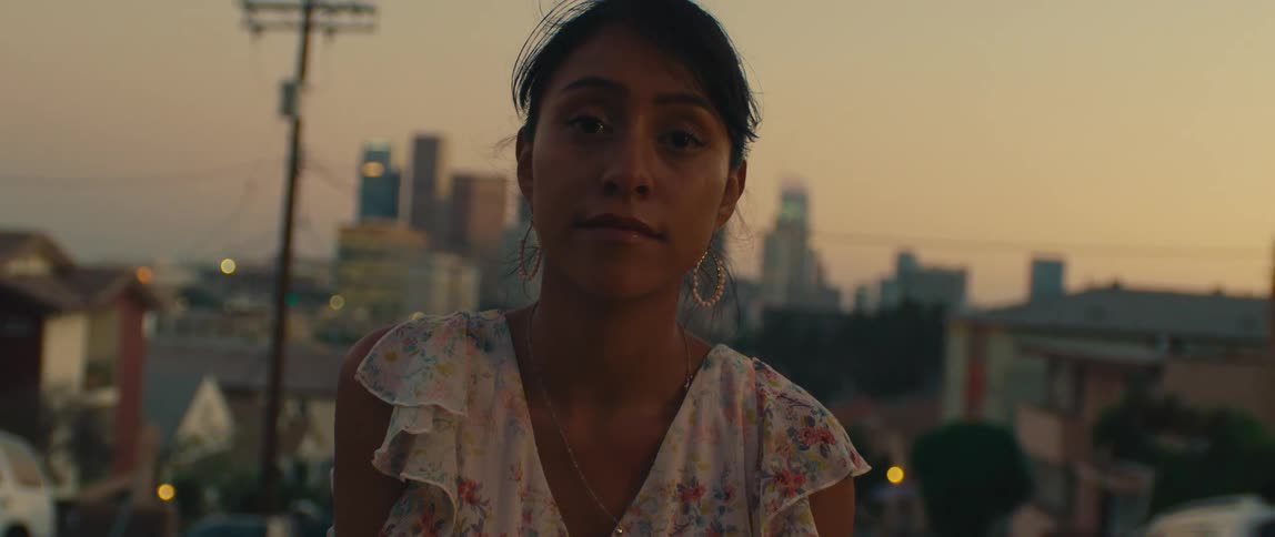 A woman with dark hair and a serious expression is shown in a medium shot looking directly at the camera she is standing on a residential street with a city skyline visible in the blurred background suggesting a view from a hill or a neighborhood near