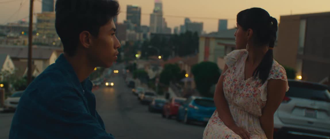 The video features a young couple looking out over a city at dusk the camera is focused on the man in the foreground wearing a dark blue shirt and a woman in a white floralpatterned dress sitting beside him in the background there is a street desce