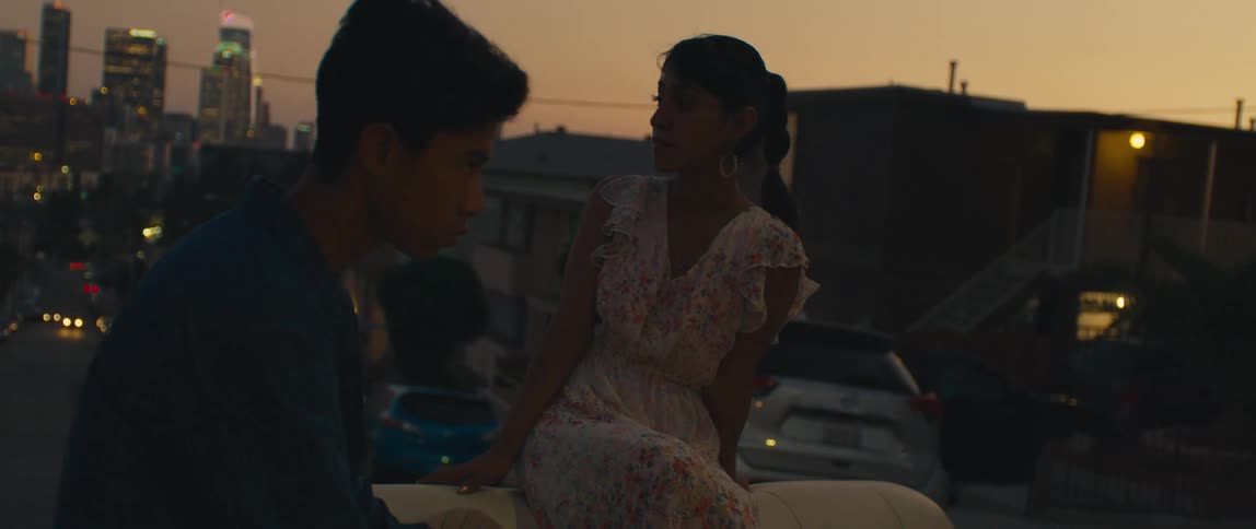 Couple talks sitting on a car at dusk with city in the background