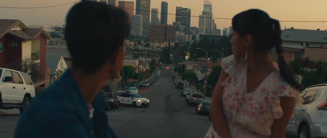 Young man in a blue shirt and a woman in a floral dress are on a steep street with city buildings in the background at sunset