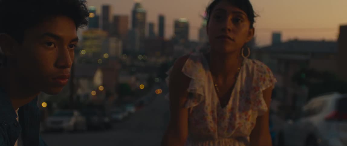 A sad looking young man and woman sit together at dusk on a residential street with the los angeles skyline glowing in the background