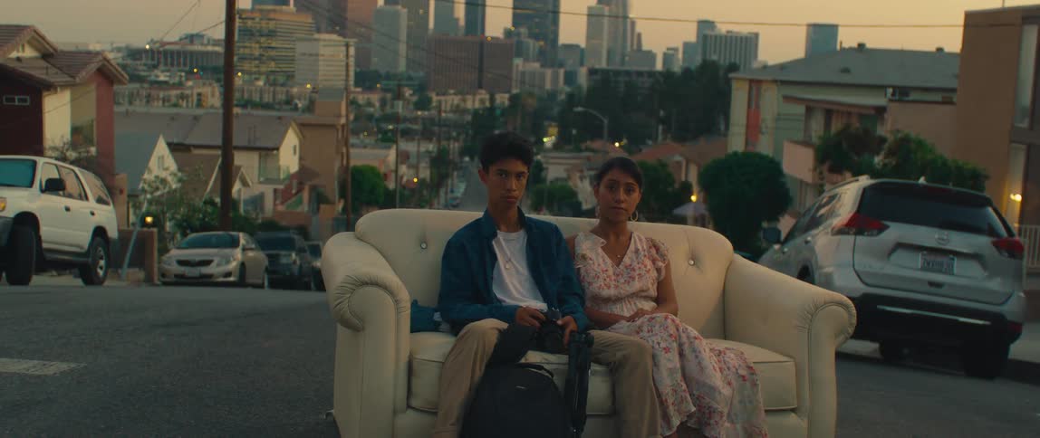Two young people sitting on a couch in the middle of a street with the city in the background
