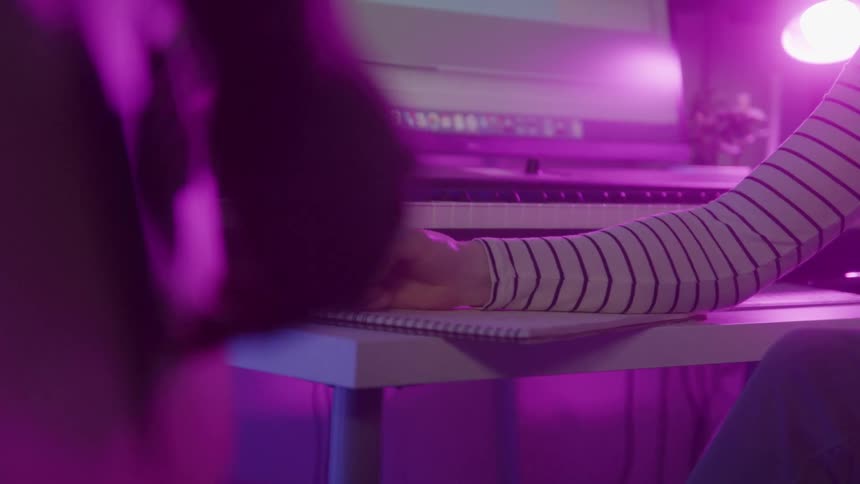 Hand writes in notebook in front of musical keyboard lit by purple light