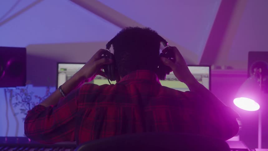 A person with dark curly hair is shown from behind sitting at a desk with a computer monitor the screen displays audio tracks suggesting they are working on music or audio production