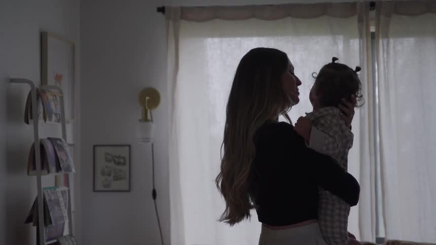 A mother gently holds a baby in a softly lit room near a window