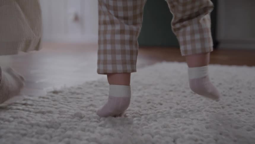 Child in checkered pajamas walks on wooden floor beside an adult