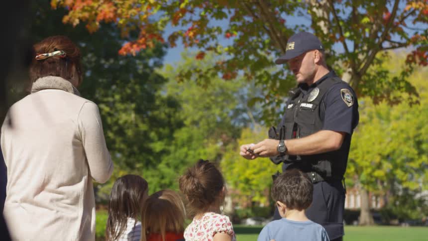 Uniformed police officer hands something to a woman while children watch in a sunny park