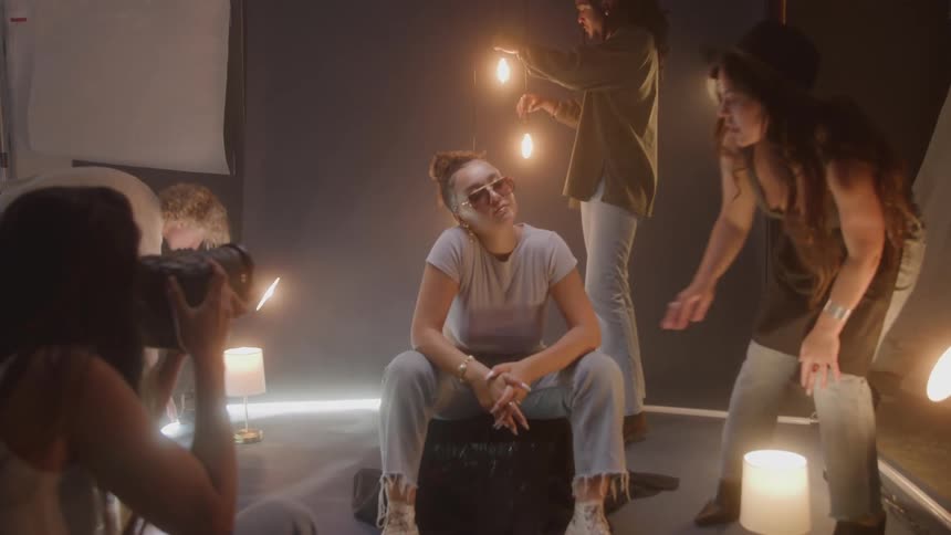 Model wearing sunglasses sitting in a studio surrounded by people and being photographed