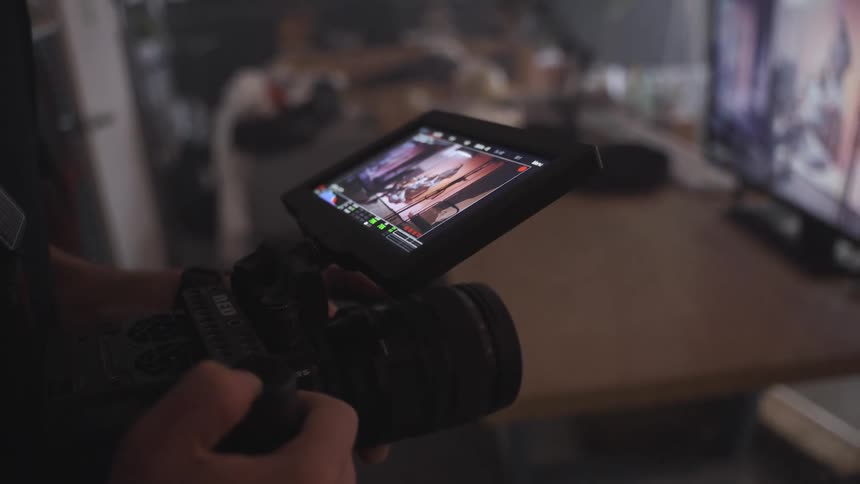 A person is holding a professional video camera with a live view screen the screen shows a film scene with a character in what appears to be a chaotic or actionfilled setting